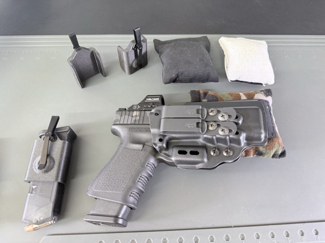 AIWB holster meaning shown in a real setup — a Kydex appendix holster holding a red-dot-equipped pistol beside a multicam Cloudster Pillow wedge, a spare-magazine carrier, and several holster wedges